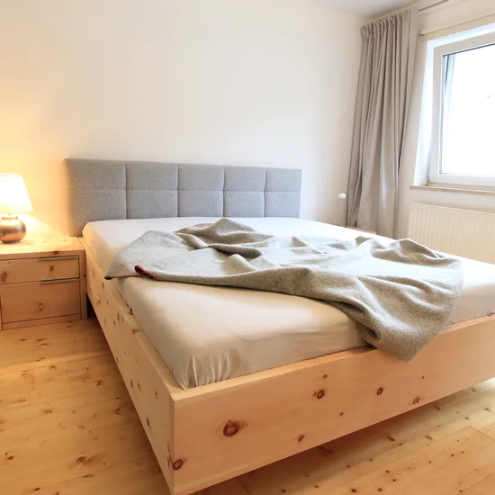 Modernes Schlafzimmer mit Holzbett, grauem Kopfteil, Nachttischlampe und Vorhängen am Fenster