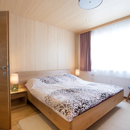 Modernes Schlafzimmer mit Holzbett, gemustertem Bettbezug und natürlichem Licht durch ein Fenster