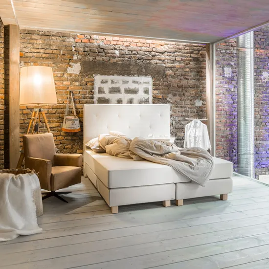 Modernes Schlafzimmer mit Boxspringbett vor rustikaler Backsteinwand und gemütlichem Sessel