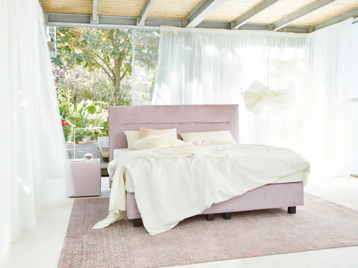 Modernes Schlafzimmer mit rosa Boxspringbett, großen Fenstern und Blick in den grünen Garten