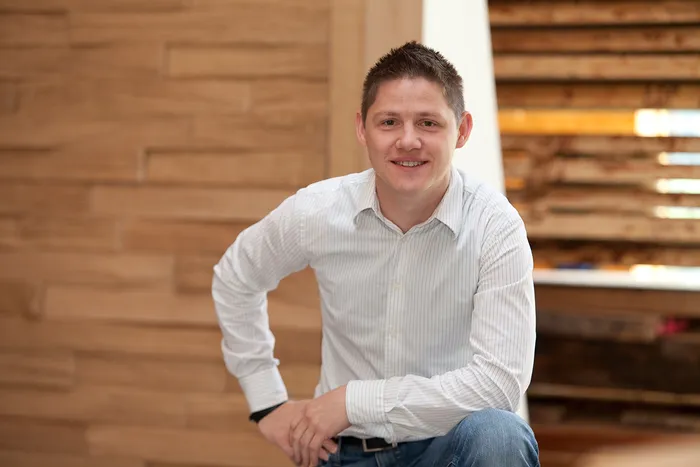 Junger Mann in weißem Hemd und Jeans lächelt vor moderner Holzinnenwand