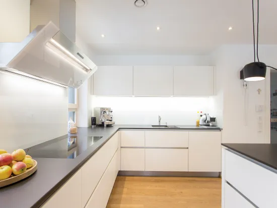 Moderne weiße Küche mit Induktionskochfeld, Dunstabzugshaube und Obstschale auf dunkler Arbeitsplatte