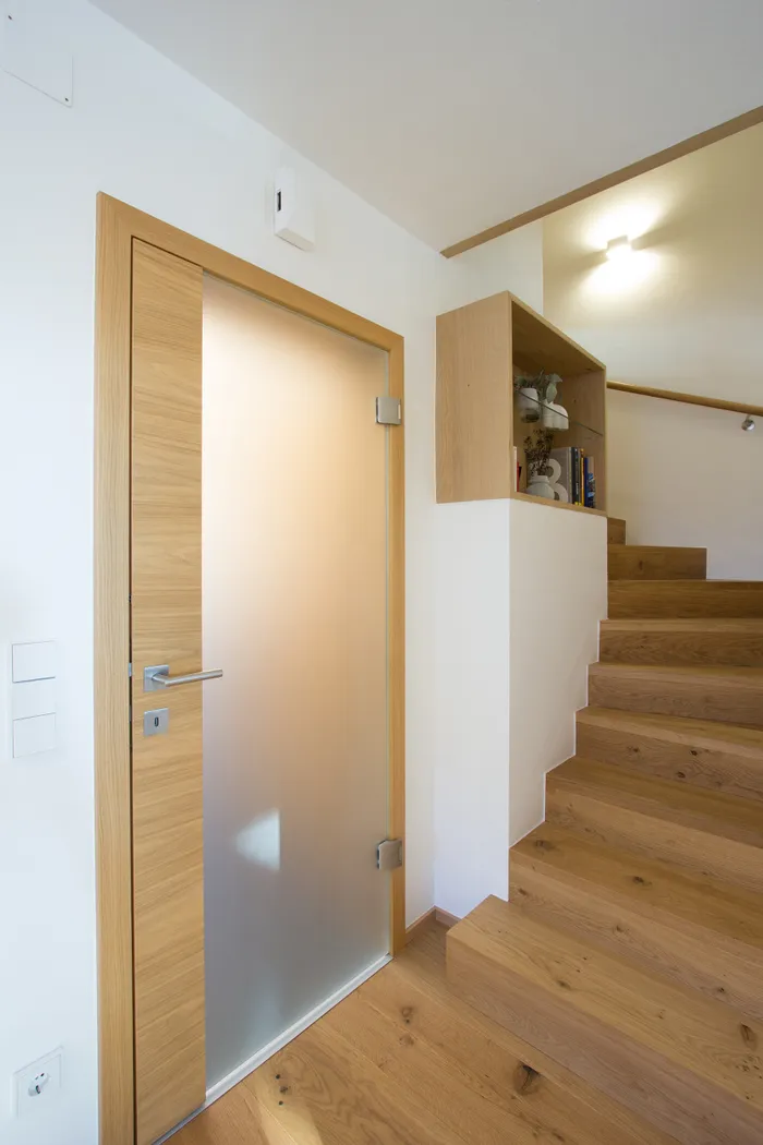 Moderne Holztreppe mit Glasinnentür und eingebautem Regal in einem hellen Flur