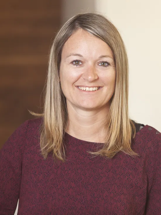 Lächelnde Frau mit glattem blondem Haar in bordeauxfarbener Bluse vor unscharfem Hintergrund