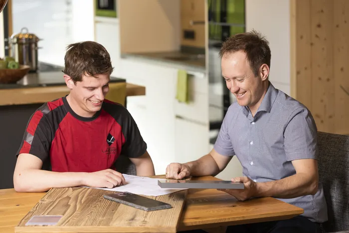 Zwei Männer besprechen Baupläne und Materialproben an einem Holztisch in moderner Küche