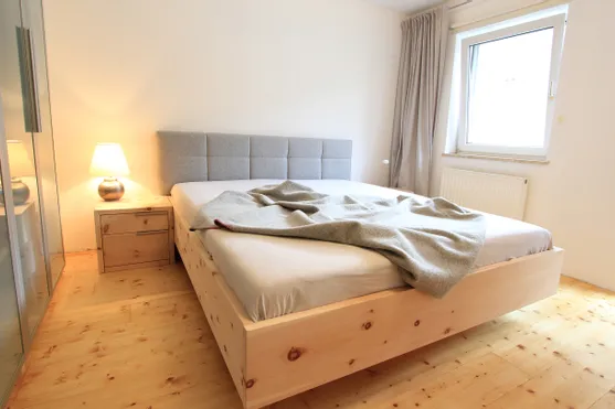 Modernes Schlafzimmer mit Holzbett, grauem Kopfteil, Nachttischlampe und Vorhängen am Fenster