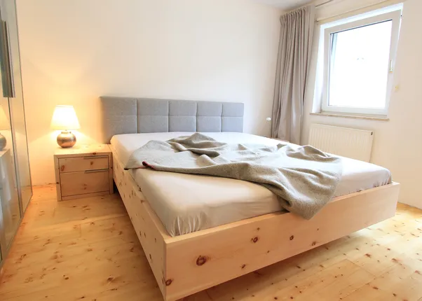 Modernes Schlafzimmer mit Holzbett, grauem Kopfteil, Nachttischlampe und Vorhängen am Fenster