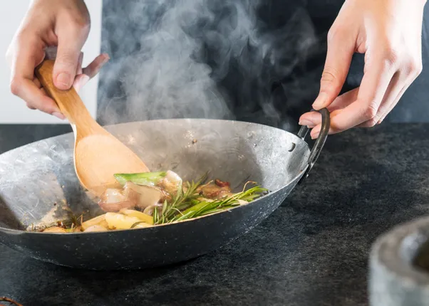 Person rührt mit Holzlöffel Gemüse und Kräuter in heißem Wok auf schwarzer Küchenarbeitsplatte