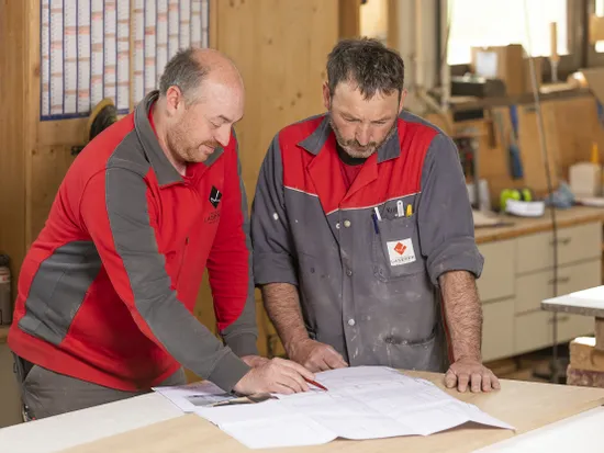 Zwei Handwerker prüfen Bauzeichnungen in einer Holzwerkstatt