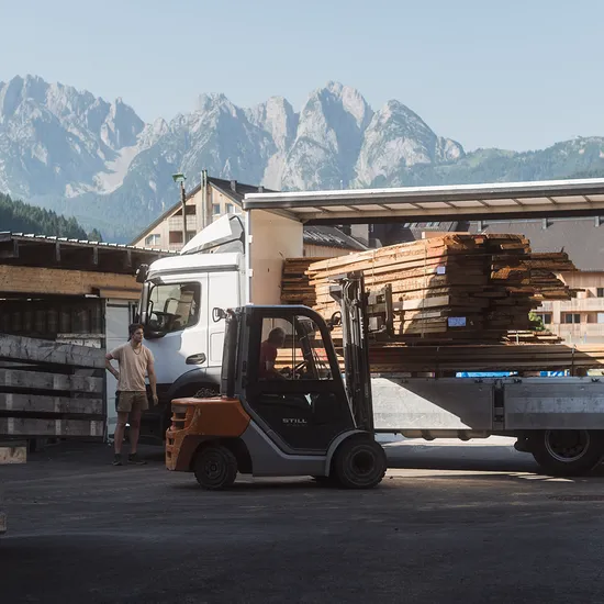 Gabelstapler lädt Holzbretter von LKW in einem Sägewerk mit Alpen im Hintergrund