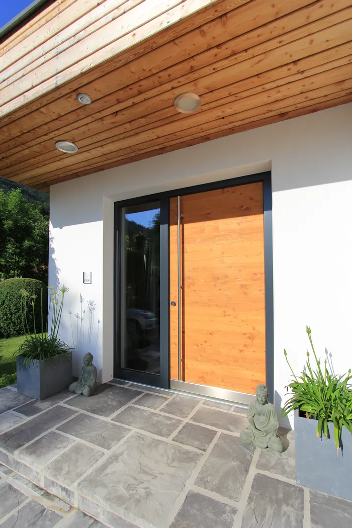 Moderne Haustür aus Holz mit Glasrahmen, flankiert von Pflanzen und Buddha-Statuen