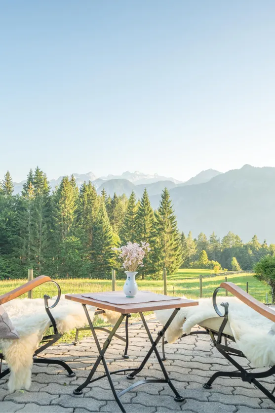 Terrasse mit zwei Stühlen und Alpenblick über grüne Wiesen und Tannen im Morgenlicht