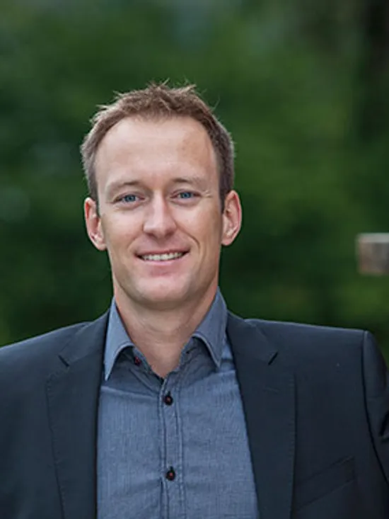 Lächelnder Mann im Anzug steht draußen neben gestapeltem Holz