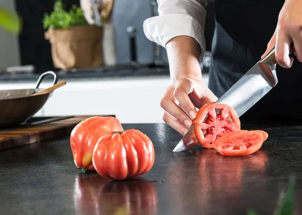 Koch schneidet reife Ochsenherztomate auf schwarzer Küchenarbeitsplatte in Scheiben
