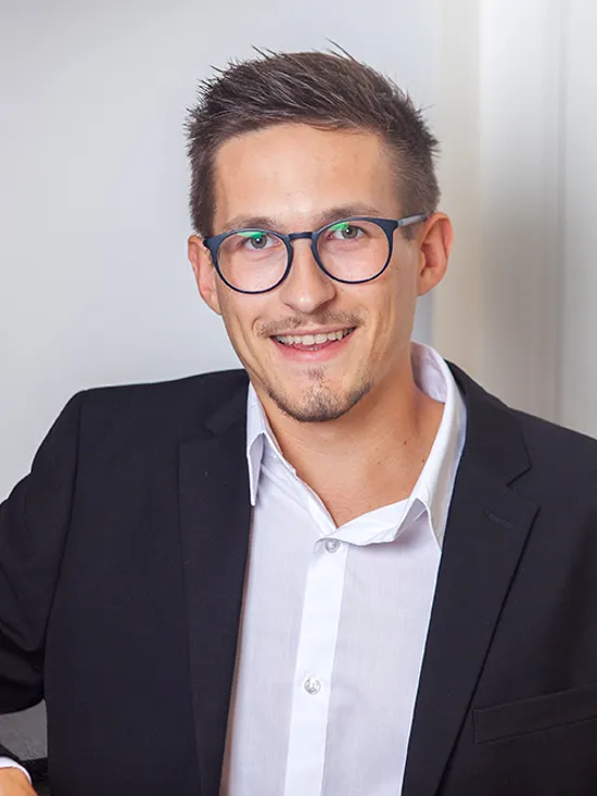 Junger Mann mit Brille im schwarzen Anzug lächelt in modernem Bürointerieur