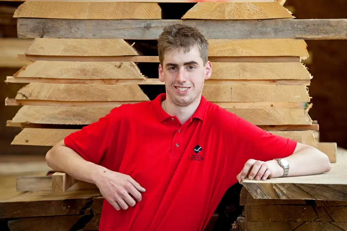 Junger Mann in rotem Poloshirt steht lächelnd vor gestapeltem Schnittholz in einer Schreinerei