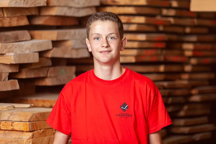 Junger Schreiner im roten T-Shirt vor gestapelten Holzbohlen in einer Tischlerei