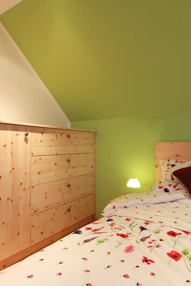 Gemütliches Schlafzimmer mit Blumenbettwäsche, grün gestrichener Wand und Möbeln aus hellem Holz