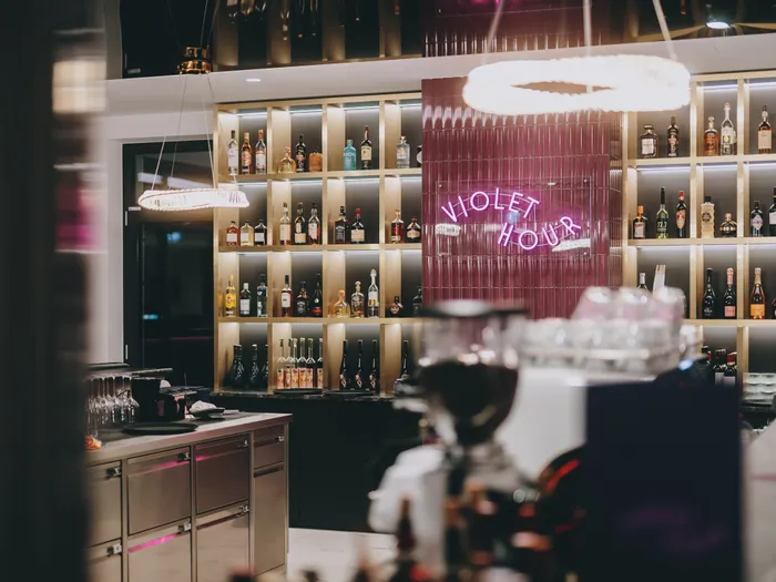 Moderne Bar mit beleuchteten Spirituosenregalen und violettem Neonzeichen Violet Hour