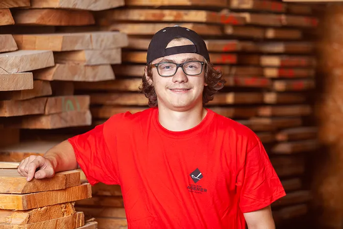 Junger Mann mit rotem T-Shirt in einem Holzlager mit gestapelten Holzbrettern