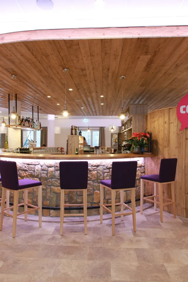 Moderne Hotelbar mit Holzdekor, lila Barhockern und Beleuchtung im COOE Hotel