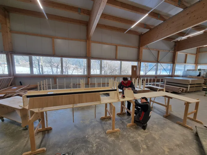 Zwei Schreiner arbeiten in einer Werkstatt an einem großen Holzmöbelstück mit Fensterblick auf den Winter