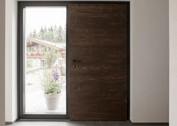 Moderne Haustür aus dunklem Holz mit seitlichem Glaselement und Blick auf ein Chalet