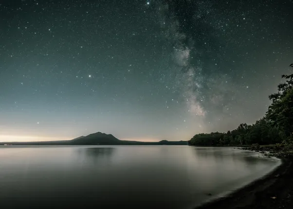 Sternenklare Nacht über ruhigem See mit Blick auf bewaldetes Ufer und Bergsilhouette