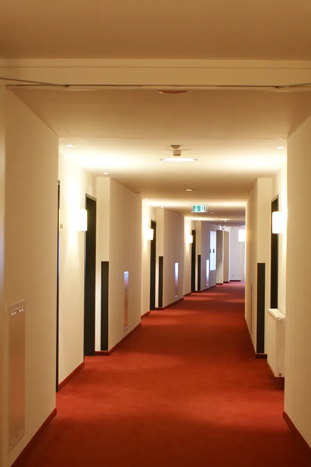 Modern hotel hallway with red carpet and white walls under warm lighting