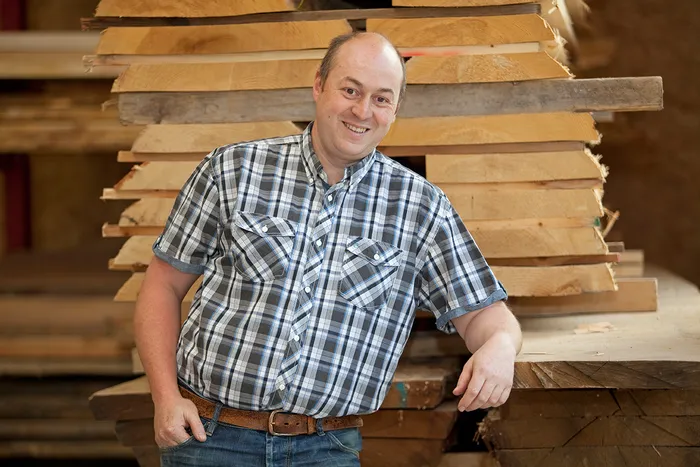 Schreiner lehnt sich lächelnd an einen Stapel Holzplatten in einer Werkstatt