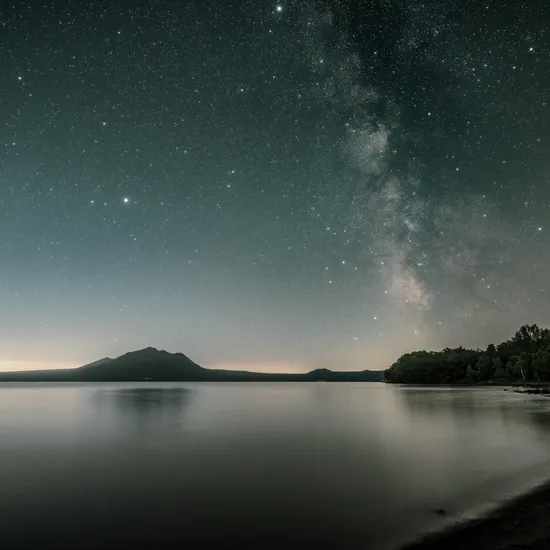 Sternenklare Nacht über ruhigem See mit Blick auf bewaldetes Ufer und Bergsilhouette