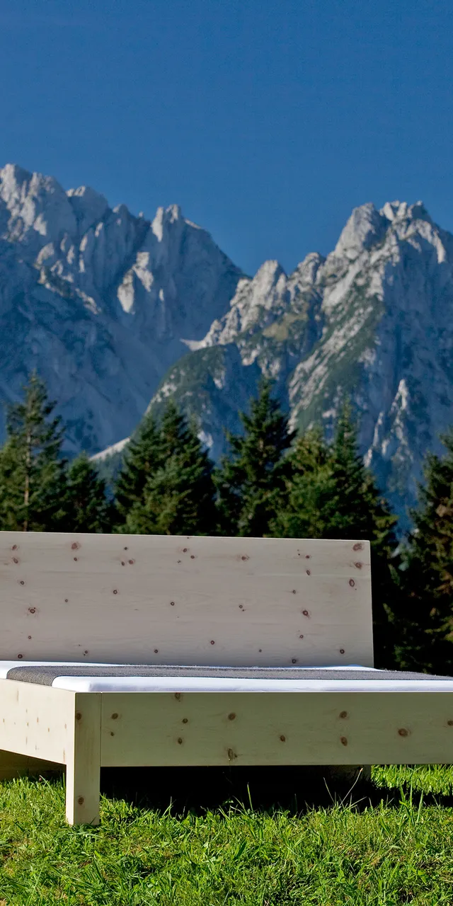 Massivholzbett aus Zirbe auf einer Wiese mit Alpenpanorama im Hintergrund