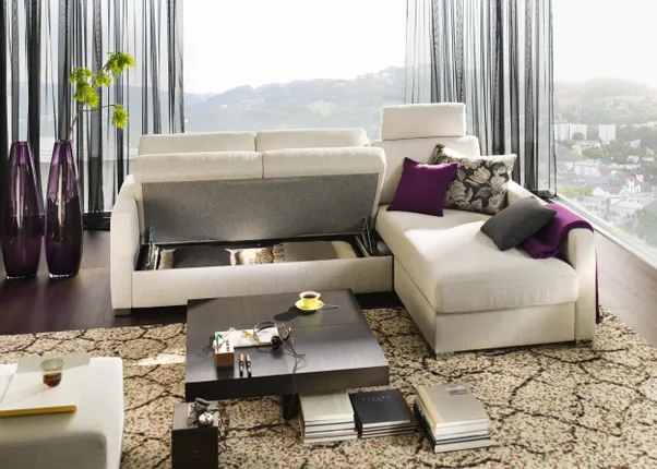 Modernes Wohnzimmer mit beigem Ecksofa, ausziehbarem Stauraum und Aussicht auf hügelige Stadtlandschaft