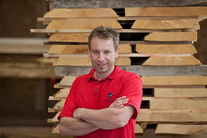 Mann in rotem Poloshirt steht vor gestapelten Holzbrettern in einer Werkstatt