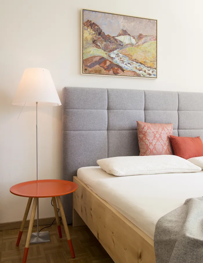 Modernes Schlafzimmer mit grauem Polsterbett, rotem Beistelltisch und Landschaftsgemälde