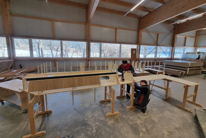 Zwei Schreiner arbeiten in einer Werkstatt an einem großen Holzmöbelstück mit Fensterblick auf den Winter