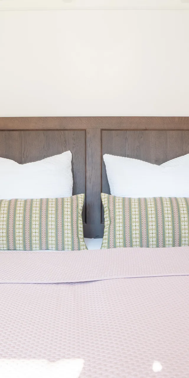 Doppelbett mit rosa Bettdecke, gemusterten Kissen und Holz-Kopfteil zwischen weißen Nachttischlampen