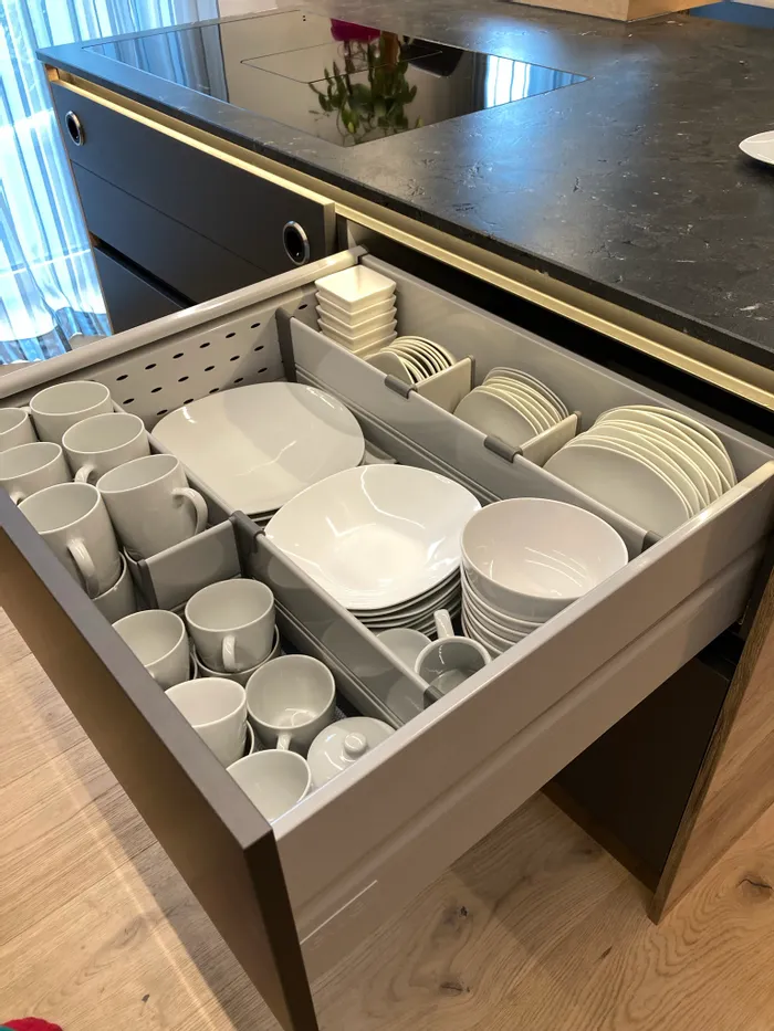 Geöffnete Küchenschublade mit ordentlich sortiertem weißem Geschirr und Tassen neben Induktionskochfeld