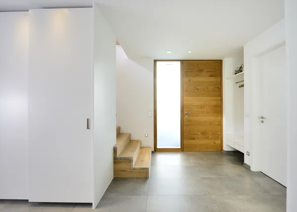 Moderner Hausflur mit Holztreppe, großem Holzeingang, weißen Wänden und grauen Bodenfliesen