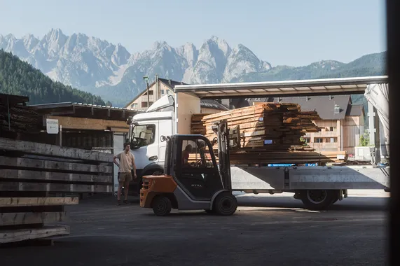 Gabelstapler lädt Holzbretter von LKW in einem Sägewerk mit Alpen im Hintergrund