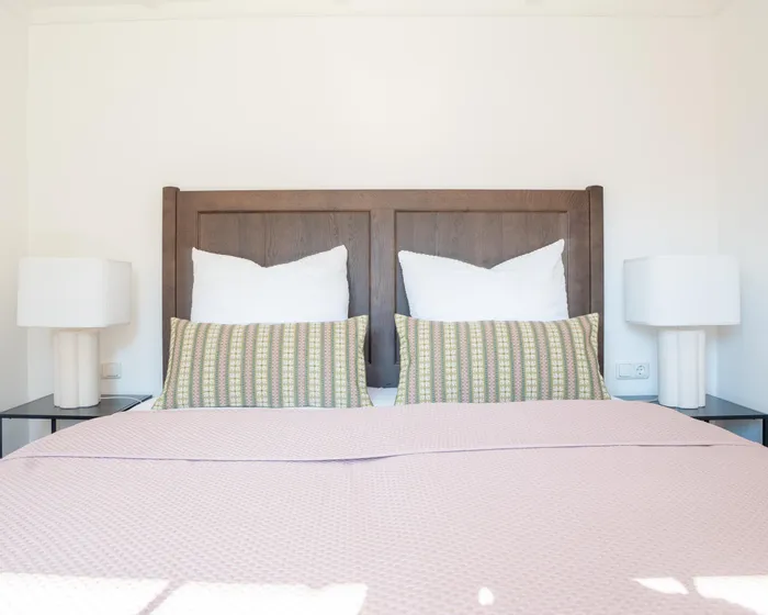 Doppelbett mit rosa Bettdecke, gemusterten Kissen und Holz-Kopfteil zwischen weißen Nachttischlampen