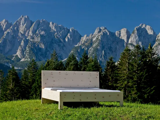 Massivholzbett aus Zirbe auf einer Wiese mit Alpenpanorama im Hintergrund