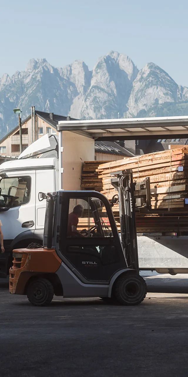 Gabelstapler lädt Holzbretter von LKW in einem Sägewerk mit Alpen im Hintergrund
