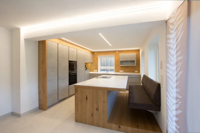 Moderne Einbauküche mit Kochinsel aus Holz, Sitzbank und indirekter LED-Beleuchtung