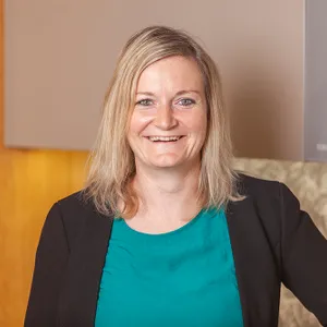 Lächelnde Frau mit blondem Haar in türkisfarbenem Oberteil und schwarzem Blazer vor modernem Hintergrund