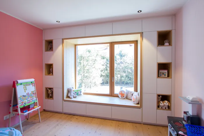 Helles Kinderzimmer mit großem Fensterplatz, Spielzeugregalen und rosa Wandfarbe