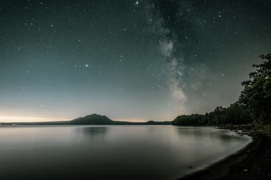 Sternenklare Nacht über ruhigem See mit Blick auf bewaldetes Ufer und Bergsilhouette