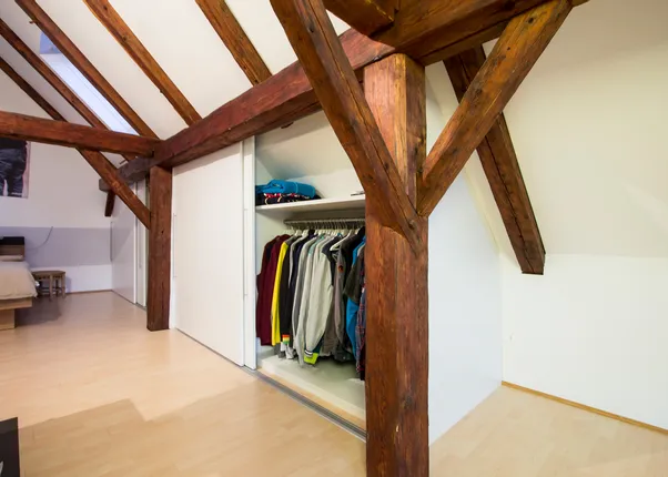 Dachgeschoss-Schlafzimmer mit sichtbaren Holzbalken und offenem Kleiderschrank mit Kleidung
