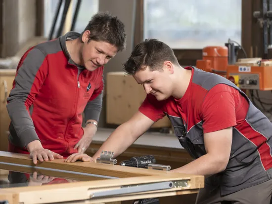 Ausbilder zeigt Auszubildendem in der Werkstatt den richtigen Umgang mit Holz und Werkzeugen