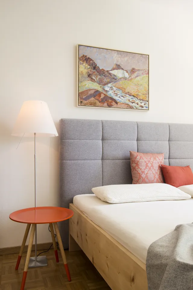 Modernes Schlafzimmer mit grauem Polsterbett, rotem Beistelltisch und Landschaftsgemälde